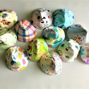 May include: A collection of 11 colorful fabric baby hats with various animal and geometric patterns. The hats are cone-shaped and made of soft, comfortable material.
