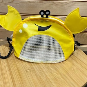 May include: A yellow crab-shaped drawstring bag with a mesh front pocket. The bag has black eyes and a black smile.