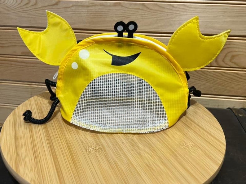 May include: A yellow crab-shaped drawstring bag with a mesh front pocket. The bag has black eyes and a black smile.