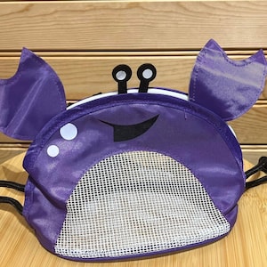 May include: Purple mesh bag shaped like a crab with a smiling face and black eyes. The bag has a drawstring closure.