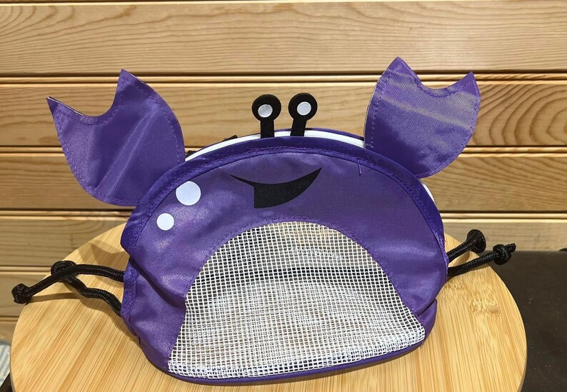 May include: Purple mesh bag shaped like a crab with a smiling face and black eyes. The bag has a drawstring closure.