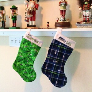 May include: Two Christmas stockings with a green and white Seahawks pattern and a blue and white plaid Seahawks pattern. The stockings are hung on a white shelf with a white trim. The stockings are personalized with the names "Holly & Krystal" and "988".