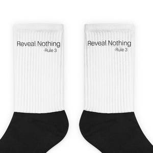 May include: A pair of white ribbed socks with black heels and toes. The text "Reveal Nothing - Rule 3" is printed in black on the upper part of each sock. The socks are displayed against a plain background.