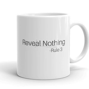 May include: White ceramic mug with a curved handle. The mug features the text "Reveal Nothing - Rule 3" in black, with a line design underneath the words. The mug is set against a white background.