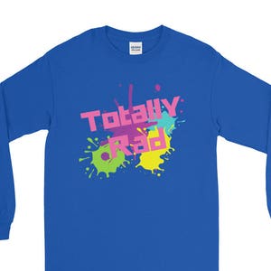May include: A royal blue long-sleeve shirt with the words "Totally Rad" in pink and yellow, with colorful paint splatters. The shirt is a solid color with a crew neck.