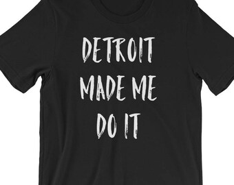 May include: Black t-shirt with the white text "DETROIT MADE ME DO IT" in a bold, handwritten style. The shirt is a short-sleeved crew neck, likely made of cotton or a similar fabric. The text is centered on the front.