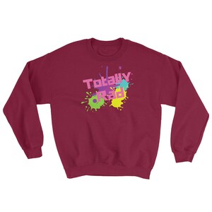 May include: A burgundy sweatshirt with the words "Totally Rad" in a colorful, 80s-style font. The text is overlaid on a splatter design in shades of pink, green, yellow, and blue. The sweatshirt has a crew neck and long sleeves.