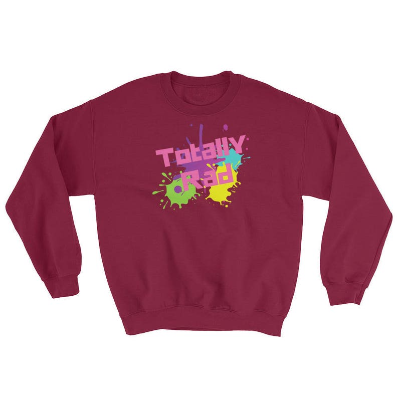 May include: A burgundy sweatshirt with the words "Totally Rad" in a colorful, 80s-style font. The text is overlaid on a splatter design in shades of pink, green, yellow, and blue. The sweatshirt has a crew neck and long sleeves.