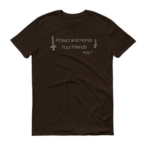 May include: A dark brown t-shirt with the text "Protect and Honor Your Friends - Rule 1" in white. The design includes two stylized sword illustrations, one on each side of the text. The shirt is a crew neck style.