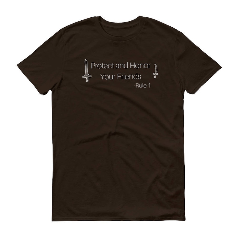 May include: A dark brown t-shirt with the text "Protect and Honor Your Friends - Rule 1" in white. The design includes two stylized sword illustrations, one on each side of the text. The shirt is a crew neck style.