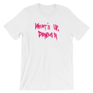 Puede incluir: Camiseta blanca con la frase "WHAT'S UP, DANGER" en letras rosas goteantes. La camiseta tiene cuello redondo y mangas cortas. El texto est&aacute; en una fuente audaz de estilo grafiti.