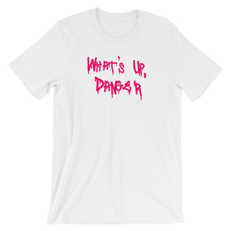 Puede incluir: Camiseta blanca con la frase "WHAT'S UP, DANGER" en letras rosas goteantes. La camiseta tiene cuello redondo y mangas cortas. El texto est&aacute; en una fuente audaz de estilo grafiti.
