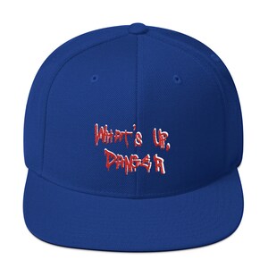 Op de afbeelding: Een koningsblauwe baseballpet met de geborduurde tekst "What's Up, Danger" in rood en wit. De pet heeft een platte klep en een gestructureerde pasvorm, geschikt voor casual wear.