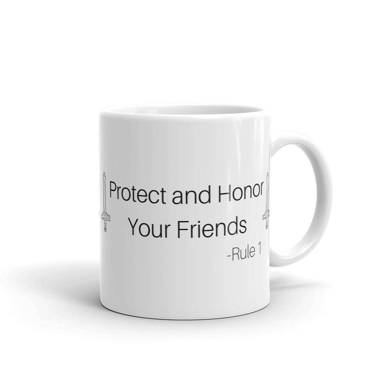 Pu&ograve; includere: Tazza in ceramica bianca con la scritta "Protect and Honor Your Friends - Rule 1" in nero. Il design include illustrazioni di spade su entrambi i lati del testo. La tazza ha un manico curvo.