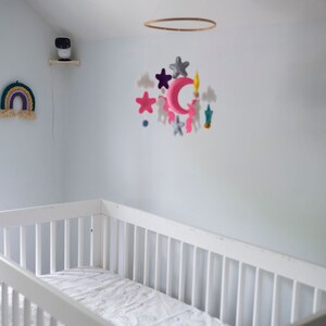 Op de afbeelding: Een witte wieg met een matras in een kinderkamer. Boven de wieg hangt een mobiel met roze, grijze en blauwe vilten vormen. Twee regenboog wandkleden en een beveiligingscamera zijn ook zichtbaar.