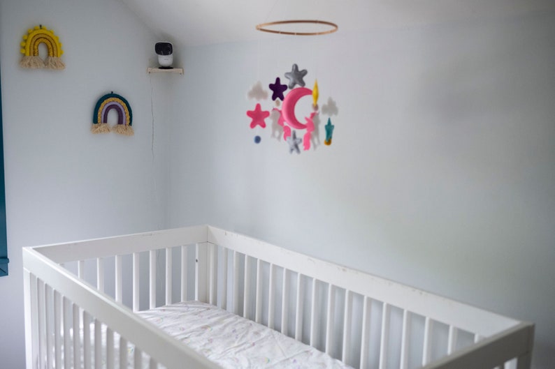Op de afbeelding: Een witte wieg met een matras in een kinderkamer. Boven de wieg hangt een mobiel met roze, grijze en blauwe vilten vormen. Twee regenboog wandkleden en een beveiligingscamera zijn ook zichtbaar.