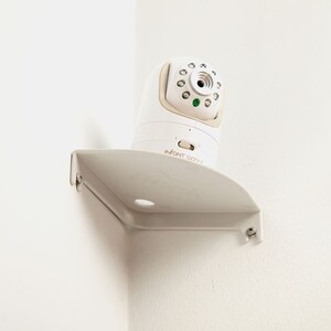 May include: A white baby monitor with a camera and a gray mounting bracket is mounted on a white wall. The monitor has the text "INFANT OPTIX" on the front.