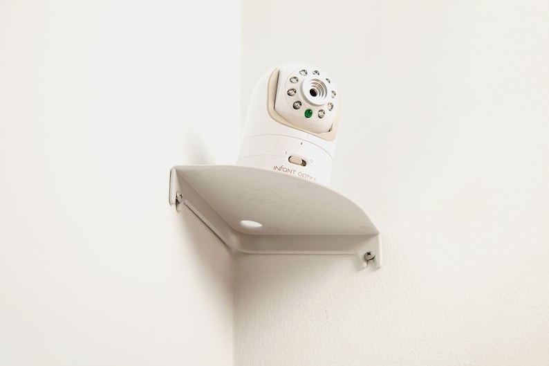 May include: A white baby monitor with a camera and a gray mounting bracket is mounted on a white wall. The monitor has the text "INFANT OPTIX" on the front.