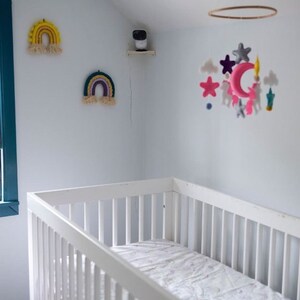 May include: A white crib with a patterned mattress pad is in a nursery. Two rainbow wall hangings and a mobile with stars, clouds, and unicorns decorate the room. A security camera is mounted on a shelf.