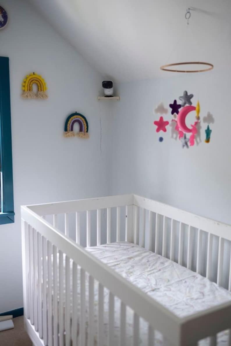 May include: A white crib with a patterned mattress pad is in a nursery. Two rainbow wall hangings and a mobile with stars, clouds, and unicorns decorate the room. A security camera is mounted on a shelf.