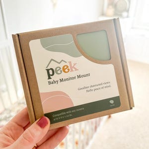 May include: A brown cardboard box labeled "peek Baby Monitor Mount" with the text "Goodbye obstructed views. Hello peace of mind." The box has a sage green and pink design and states it is compatible with any camera. Dimensions are 4.75 x 6 x 1.12 in.