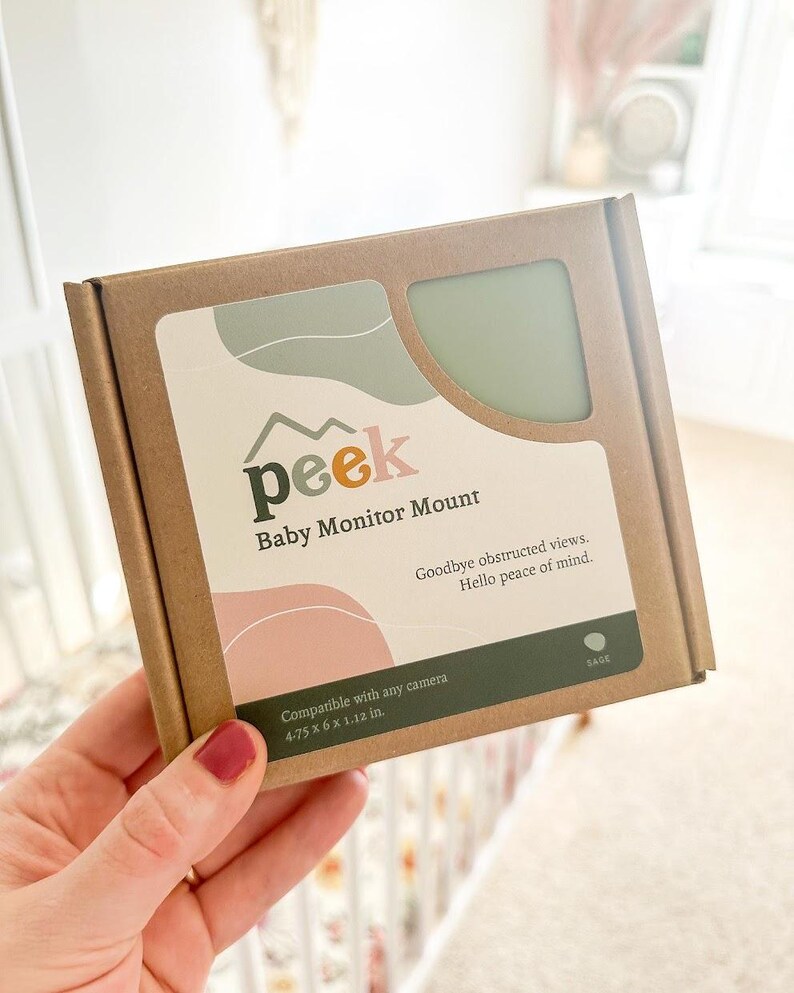 May include: A brown cardboard box labeled "peek Baby Monitor Mount" with the text "Goodbye obstructed views. Hello peace of mind." The box has a sage green and pink design and states it is compatible with any camera. Dimensions are 4.75 x 6 x 1.12 in.