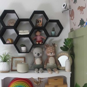 May include: A white bookshelf with a rainbow wooden toy, stuffed animals, and decorative items. Black hexagon shelves hold books, a doll, and a small cactus. The room has a dinosaur-themed wallpaper and a small mirror.
