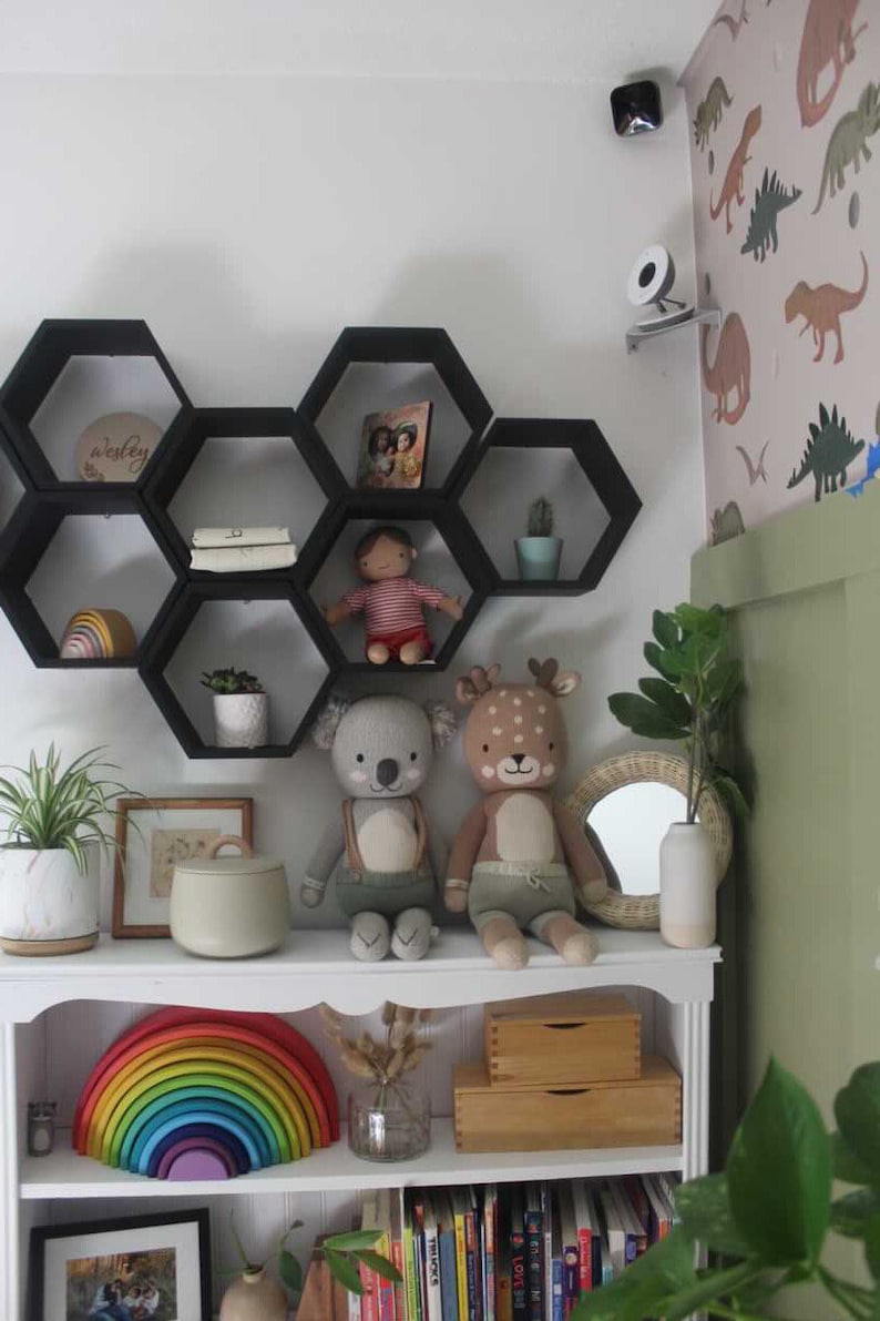 May include: A white bookshelf with a rainbow wooden toy, stuffed animals, and decorative items. Black hexagon shelves hold books, a doll, and a small cactus. The room has a dinosaur-themed wallpaper and a small mirror.