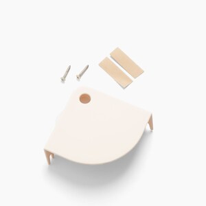 Op de afbeelding: Een lichtbeige hoekplank met twee schroeven en twee plakstrips. De plank heeft een afgeronde vorm met een gat en twee kleine pootjes. De items staan op een witte achtergrond.