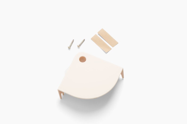 Op de afbeelding: Een lichtbeige hoekplank met twee schroeven en twee plakstrips. De plank heeft een afgeronde vorm met een gat en twee kleine pootjes. De items staan op een witte achtergrond.