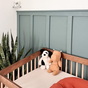 Op de afbeelding: Een houten babybedje met een lichtbeige hoeslaken, twee knuffels en een oranje kussen. Een babyfoon is aan de muur boven het bedje bevestigd. De muur achter het bedje is in een gedempte blauwgroene kleur geschilderd.