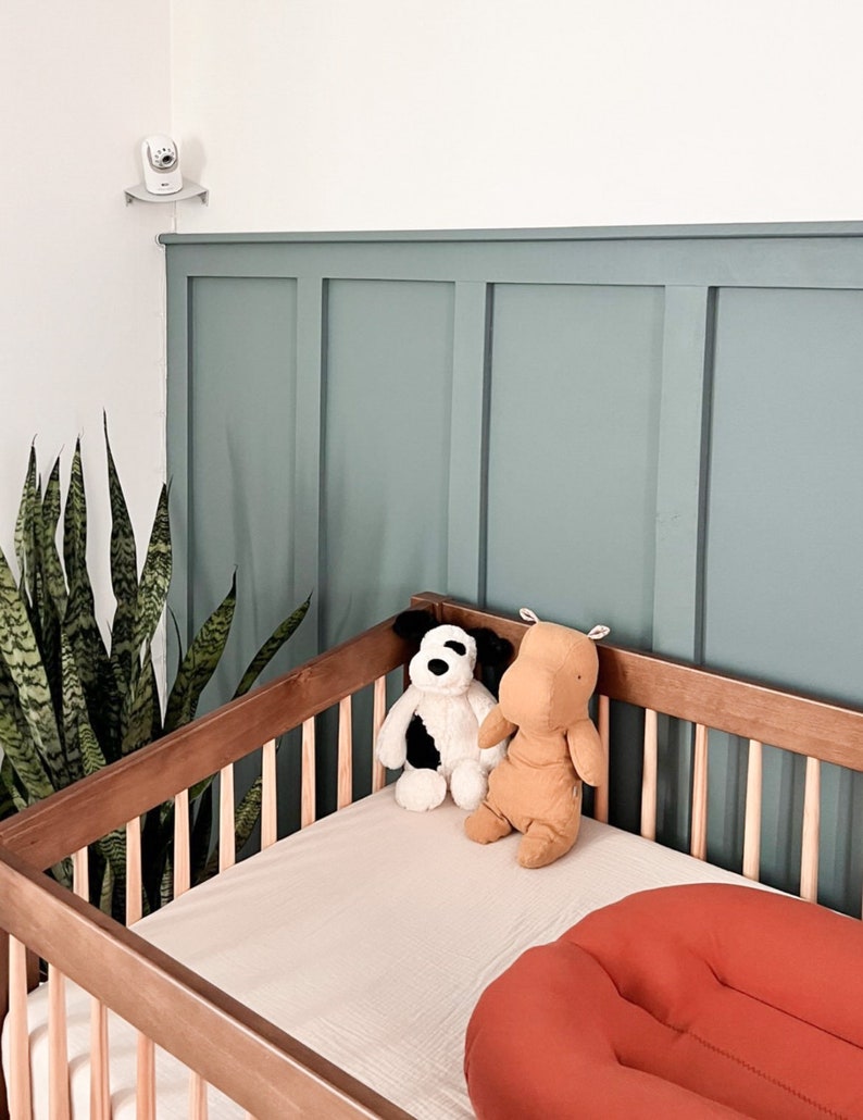 Op de afbeelding: Een houten babybedje met een lichtbeige hoeslaken, twee knuffels en een oranje kussen. Een babyfoon is aan de muur boven het bedje bevestigd. De muur achter het bedje is in een gedempte blauwgroene kleur geschilderd.