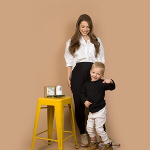 May include: A yellow metal stool with a square seat and four legs. A woman in a white shirt and black pants stands next to a young child in a black shirt and white pants. A small box and a white object sit on the stool.