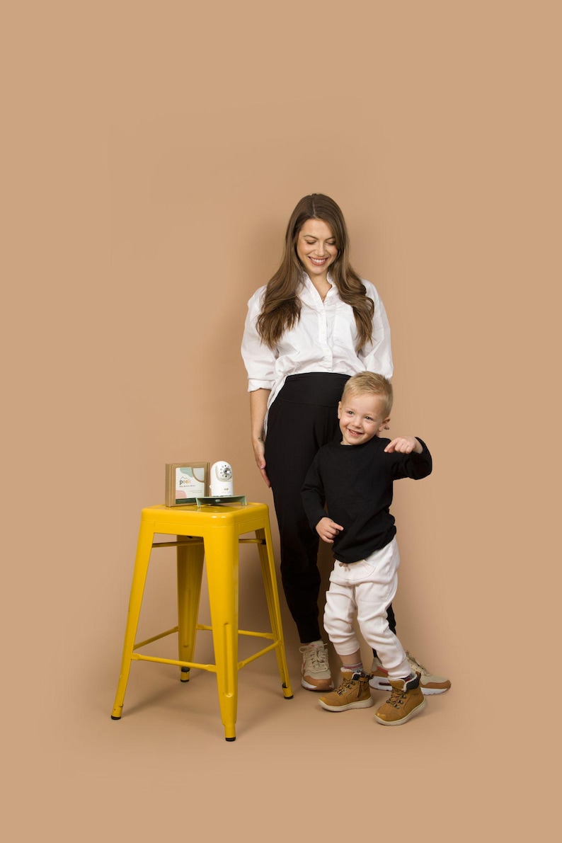 May include: A yellow metal stool with a square seat and four legs. A woman in a white shirt and black pants stands next to a young child in a black shirt and white pants. A small box and a white object sit on the stool.
