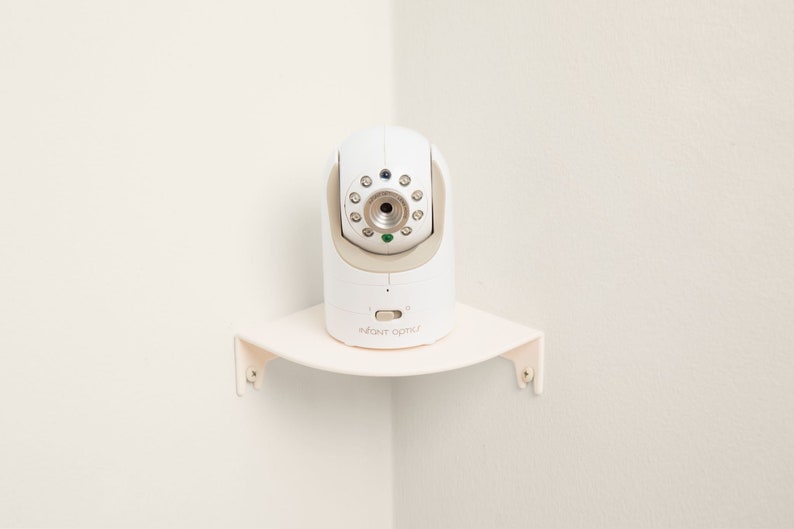 Op de afbeelding: Een witte babyfoon met een cameralens en meerdere lampjes, gemonteerd op een hoekplank. De monitor heeft de tekst "INFANT OPTICS" en een aan/uit-knop. De plank is lichtcr&egrave;me van kleur en is in een hoek gemonteerd.