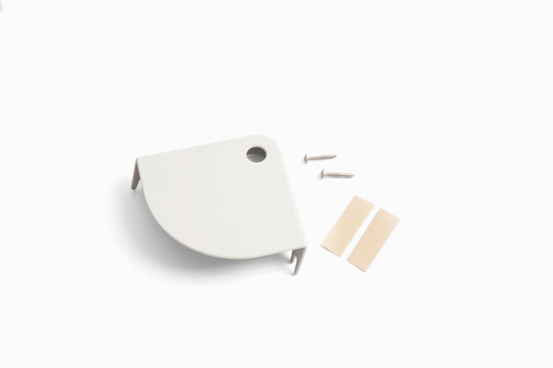 May include: A white, corner-shaped shelf bracket with a pre-drilled hole, two silver screws, and two beige adhesive strips. The bracket has a curved front edge and two small support tabs.