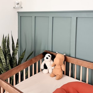 May include: A wooden baby crib with a light beige fitted sheet, two stuffed animals, and an orange pillow. A baby monitor is mounted on the wall above the crib. The wall behind the crib is painted a muted teal color.