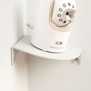 Op de afbeelding: Een witte babyfoon met een beige accent, gemonteerd op een witte plank. De monitor heeft een lens en meerdere lampjes. De merknaam "INFANT OPTIC" staat op de voorkant.