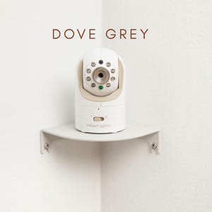 May include: A white and beige baby monitor with the text "INFANT OPTICS" is mounted on a dove grey corner shelf. The monitor has a camera lens and several small lights. The words "DOVE GREY" are above the monitor.