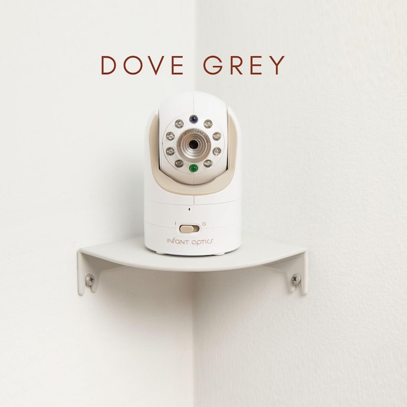 May include: A white and beige baby monitor with the text "INFANT OPTICS" is mounted on a dove grey corner shelf. The monitor has a camera lens and several small lights. The words "DOVE GREY" are above the monitor.