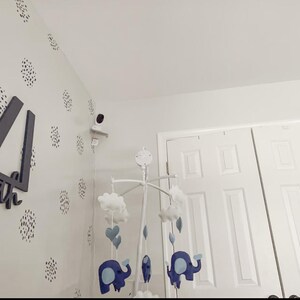 May include: A baby mobile with blue elephants, hearts, and clouds hangs in a nursery. The mobile is suspended from a white frame. The wall is decorated with black and white polka dots and a security camera.