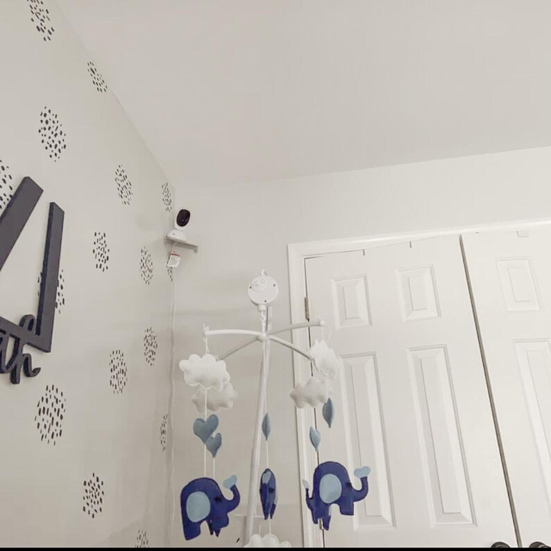 May include: A baby mobile with blue elephants, hearts, and clouds hangs in a nursery. The mobile is suspended from a white frame. The wall is decorated with black and white polka dots and a security camera.