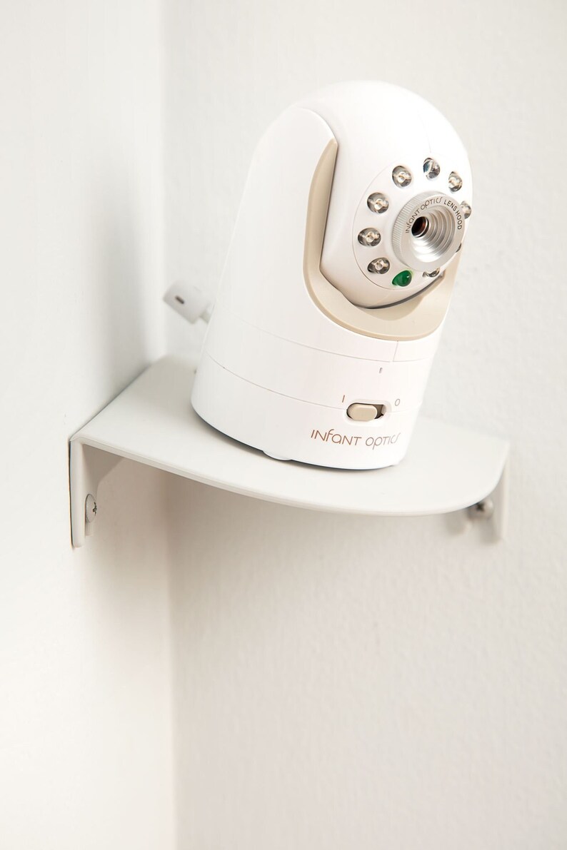May include: A white baby monitor with a beige accent, mounted on a white shelf. The monitor has a lens, several lights, and the text "INFANT OPTIC" printed on the front. The shelf is attached to a white wall.