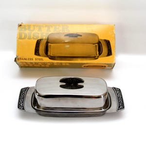 May include: A rectangular stainless steel butter dish with a polished finish, featuring a lid with a black handle and decorative black floral accents on the side handles. The original yellow cardboard box is included, labeled BUTTER DISH.