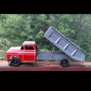 May include: A vintage-style metal toy dump truck featuring a bright red cab and a grey tilting bed. The truck has black wheels and a functional hinge mechanism that allows the rear cargo bed to be raised into an angled dumping position.