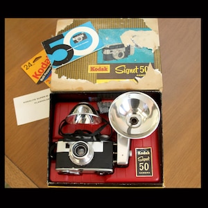 May include: A vintage Kodak Signet 50 camera set in its original box, featuring a black and silver camera body, a detachable chrome flash reflector, and a red plastic insert. The box lid displays the text Kodak Signet 50 and a large blue 50 graphic.