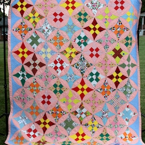 May include: A patchwork quilt featuring a repeating pattern of multi-colored fabric squares arranged in diamond shapes. The quilt is bordered by light blue fabric and accented with soft pink curved shapes that connect the colorful diamond blocks.