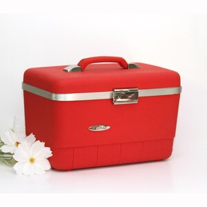 May include: A rectangular red hard-shell vanity case with a textured finish, featuring a red top handle, a silver-toned metal latch closure, and a silver trim band. The case has a small oval logo plate on the front and a structured base design.