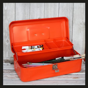 May include: A bright orange metal storage box with a hinged lid and a cantilevered tray. The interior tray holds several paint tubes, while the bottom compartment contains various paintbrushes and colored pencils. A metal latch secures the front.