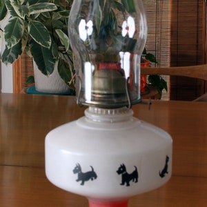 May include: A red and white oil lamp with a clear glass chimney. The lamp has a white base with black silhouettes of Scottish terriers.